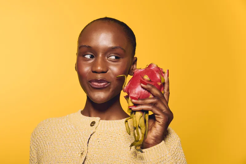 Smiling dark skinned African woman holds red dragon fruit near face on yellow background. Concept of healthy lifestyle, exotic fruit, beauty, and natural skincare with fresh tropical food.