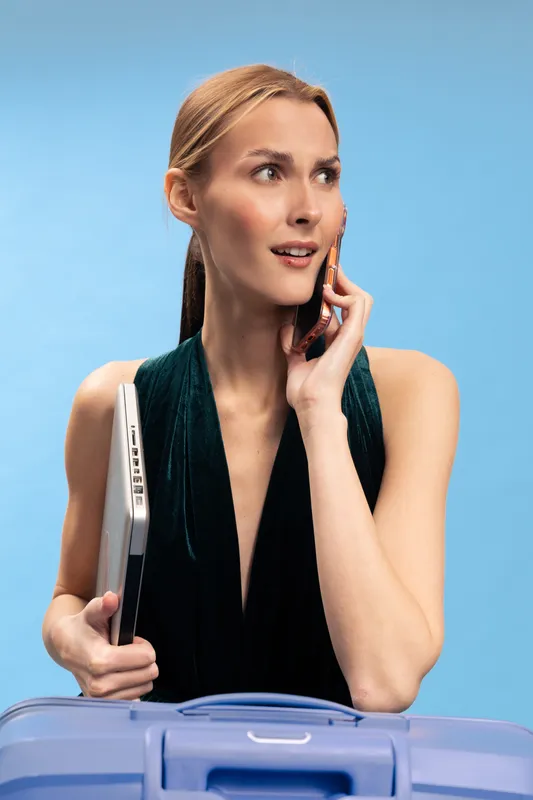 Young businesswoman holding laptop and suitcase talks on smartphone, looking away with a focused expression. Concept of travel, communication, and professional lifestyle.
