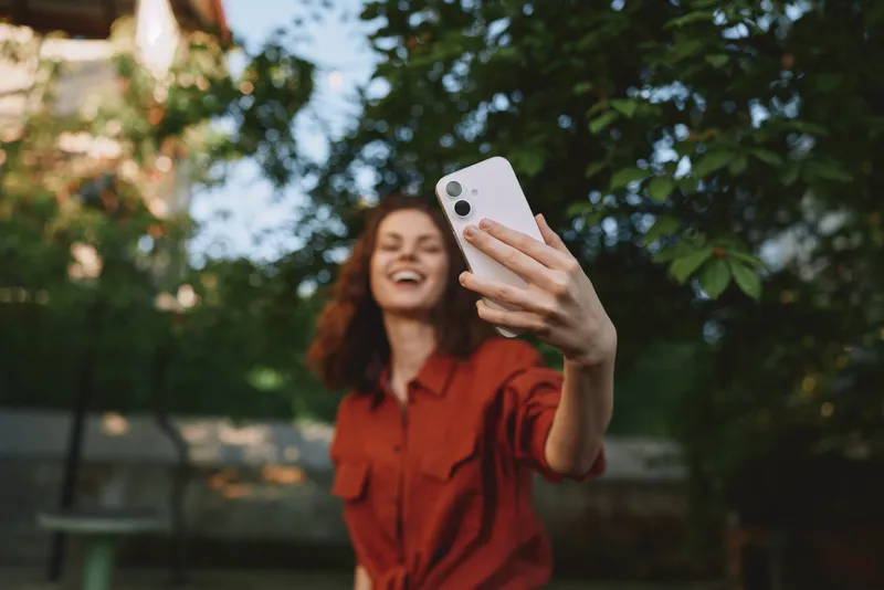 Young woman with redhead hair takes a sunny selfie outdoors, smiling in a casual red shirt against lush greenery