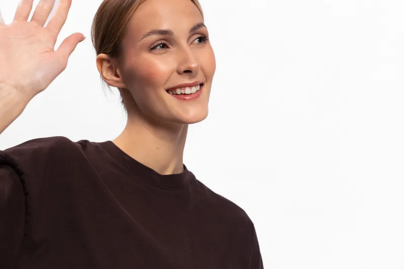 Smiling young woman in dark sweater waves hand, looking away on white background. Portrait of friendly female greeting, positive emotion, casual style and happy mood in studio setting.