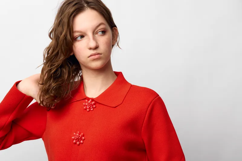 Young woman with curly hair wearing a red sweater looks away thoughtfully against a plain white background. Concept of casual fashion, contemplation, and natural beauty.