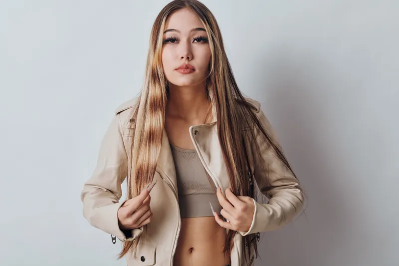 Woman with long blonde hair wearing beige jacket and crop top standing against light background looking at camera with confident expression and hands holding jacket edges.