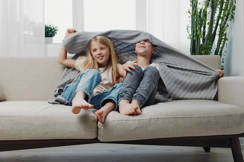 Two happy children sitting barefoot on a sofa covered with a blanket, playing and smiling in a bright modern living room with indoor plants and natural light.