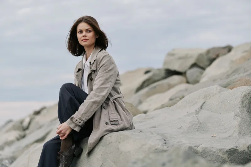 Young woman in a beige coat sits on large rocks outdoors, looking thoughtfully into the distance under an overcast sky. Concept of contemplation, nature, and autumn fashion.