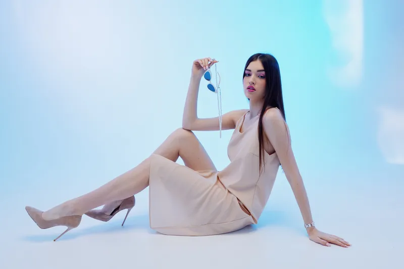 Elegant woman in a beige dress and high heels poses sitting on the floor holding sunglasses. Studio shot with soft blue lighting. Concept of beauty fashion and style.