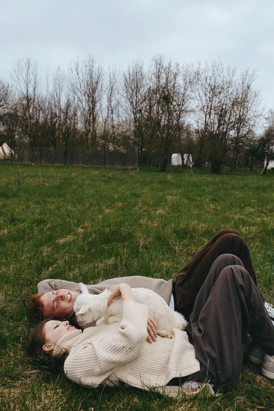 Relaxed couple lying on grass with a goat kid beside them, cozy sweaters, natural light, outdoor lifestyle moment, calm rustic mood, affectionate cuddle, peaceful countryside backdrop, candid family