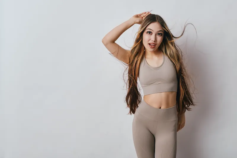 Young woman in sportswear posing with hand on head, long hair flowing, looking at camera with playful expression against white background for fitness or lifestyle.