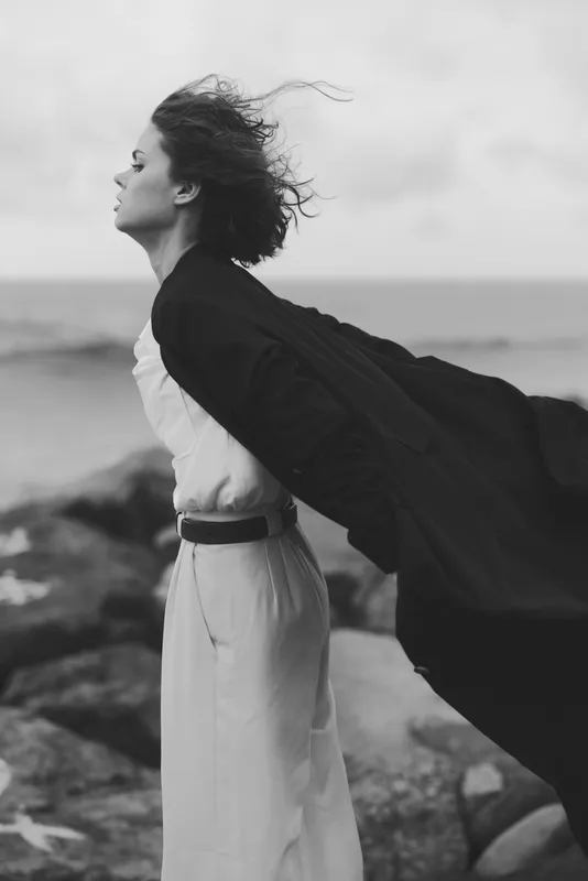 Wind sweeps a flowing cape and dress along a windswept beach, creating a dramatic fashion moment by the sea with moody tones and graceful movement.