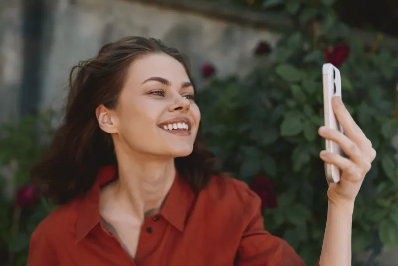 Woman lifestyle selfie in outdoor garden, smiling as she poses for a photo, casual red blouse under natural light, a cheerful candid moment