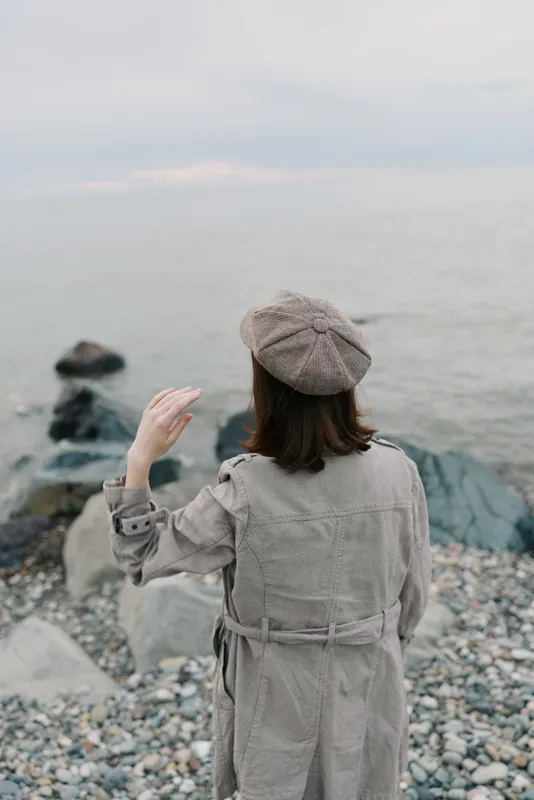 Woman in beige trench coat and patterned beret stands on rocky shore facing calm sea. Overcast sky and muted tones create quiet, reflective mood with natural coastal scenery.