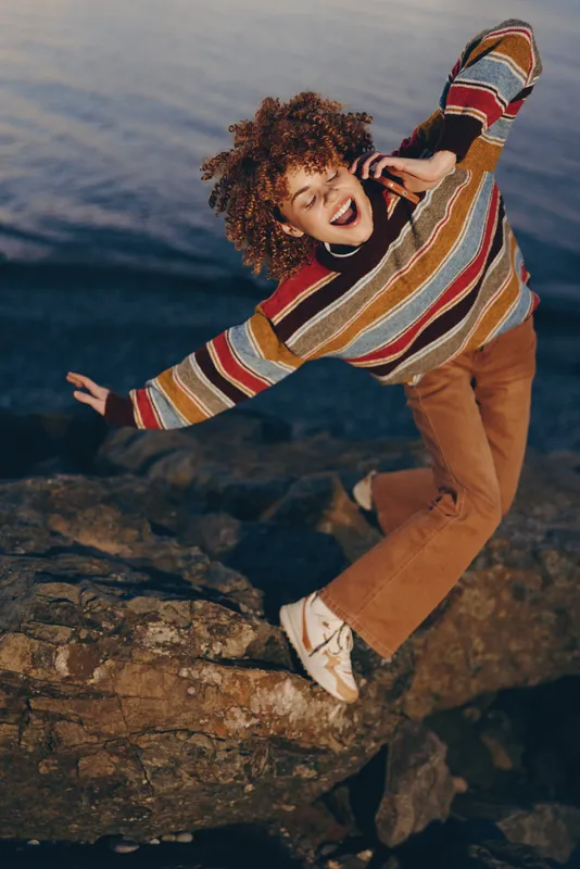 Woman with curly hair in a striped sweater and jeans climbs rocks by the seaside, lifestyle outdoor with a smile. Sunny daylight, waves, rugged stone textures and adventure feeling.