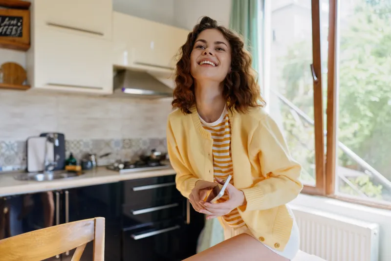 Young woman in a bright kitchen holds a phone and a snack, wearing a yellow jacket and striped shirt, smiling warmly, a casual home lifestyle moment, natural light, relaxed vibe and joy.