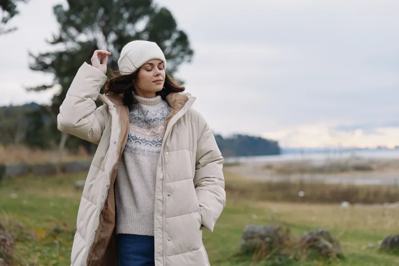 Outdoor portrait of a woman in a puffy coat and beanie in a windy countryside, calm mood, soft light, winter fashion, nature backdrop, serene atmosphere