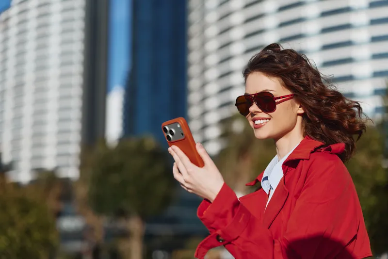 Young woman with curly hair and sunglasses smiles while using a smartphone outdoors in a city environment on a sunny day wearing a red jacket and white shirt.