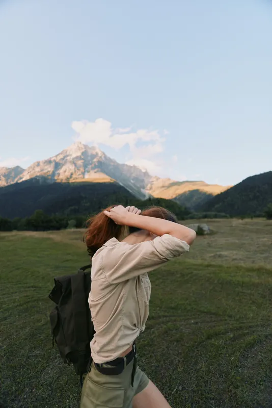 Young woman with backpack walking in green field near mountains under clear sky. Outdoor lifestyle, travel and adventure concept with natural light and casual clothing.
