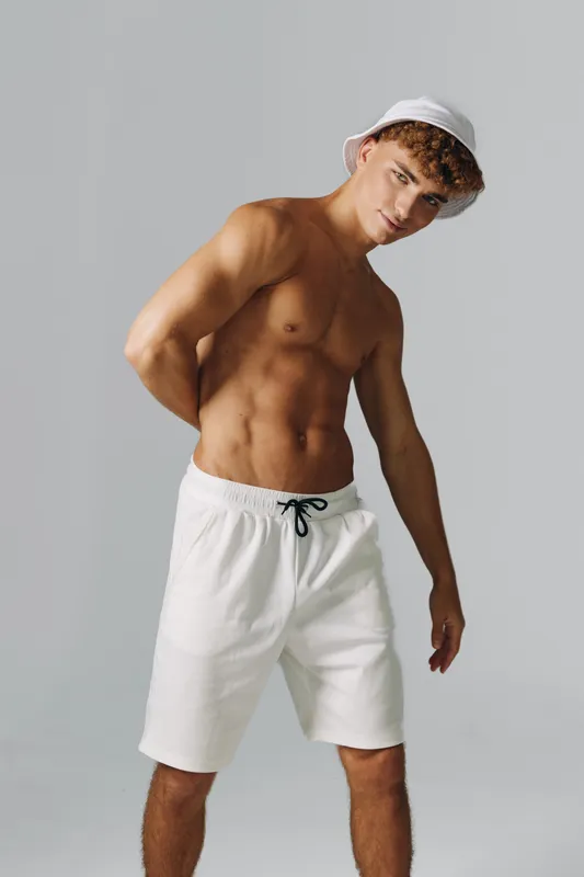 Young man with muscular body wearing white shorts and bucket hat posing against gray background. Fitness model showing toned abs and casual summer style with confident expression.