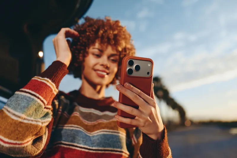 Woman lifestyle, smiling curly hair woman takes a selfie outdoors at sunset, holding a smartphone in hand, warm evening light, street background, candid moment
