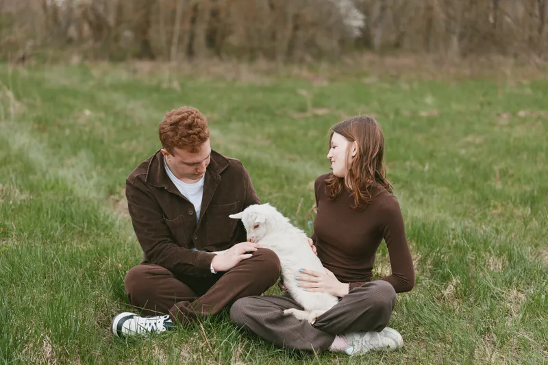 Goat kid with couple outdoors, calm interaction in a sunlit pasture, natural colors, relaxed posture, lifestyle moment capturing affection, rustic setting and gentle companionship