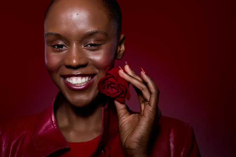 Close-up of smiling dark skinned African woman holding red rose near face. She wears red leather jacket and has natural makeup. Concept of beauty, confidence, and floral elegance.