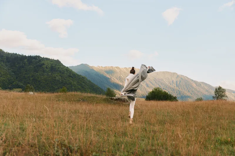 Woman lifestyle walking in grassy field with mountains in background, wrapped in blanket, enjoying nature, sunny day, casual clothing, peaceful outdoor scene