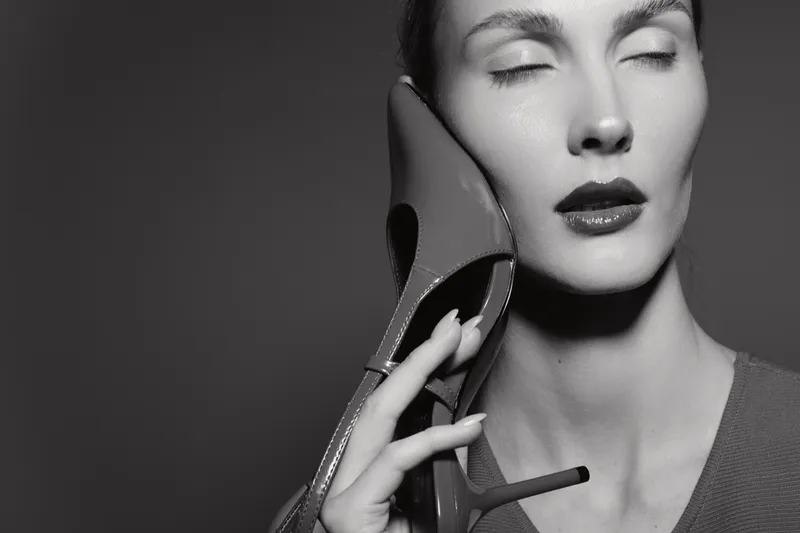 Caucasian woman with pale skin holds high heel shoe near face, eyes closed, in black and white studio portrait. Concept of fashion and elegance.