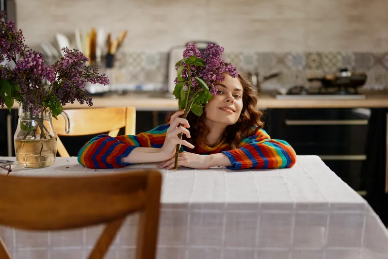 Woman in a colorful sweater rests her head on a table while holding a bouquet of flowers, smiling softly in a cozy home kitchen, portraying a warm lifestyle moment and gentle mood.