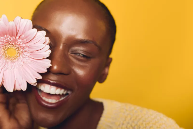 Close-up of happy African woman with dark skin holding pink flower near face, smiling with closed eyes on yellow background. Concept of beauty, wellness, and natural skincare.
