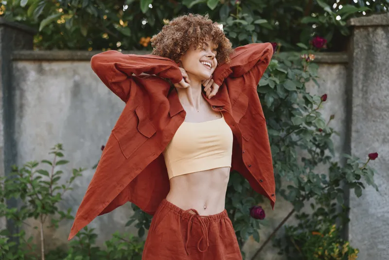 Young woman with curly hair smiles as she lifts a rust jacket over a beige crop top in an outdoor garden scene, capturing a bright, confident moment of casual fashion