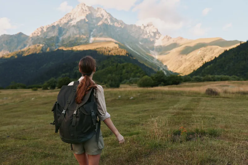 A young woman with long brown hair and a large backpack hikes across a grassy field towards a towering mountain under a partly cloudy sky.
