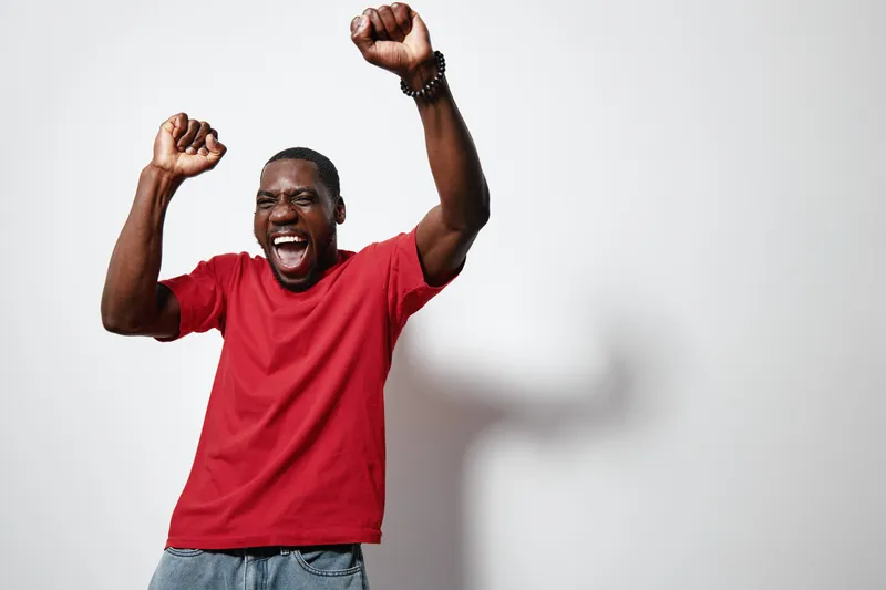 African American man with dark skin wearing red t shirt and jeans celebrating with raised hands and joyful expression on white background in studio.