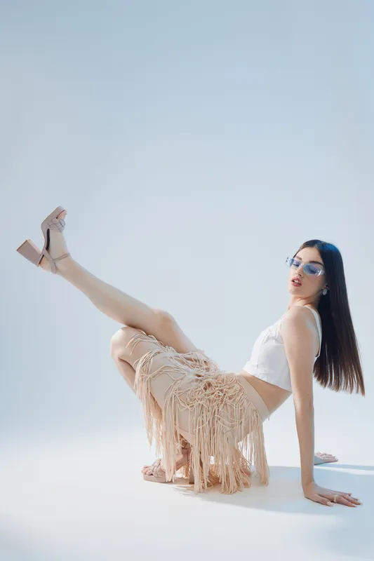 Young woman in white top and fringed skirt poses with raised leg wearing sunglasses and heels on light background. Concept of woman beauty fashion and modern style.