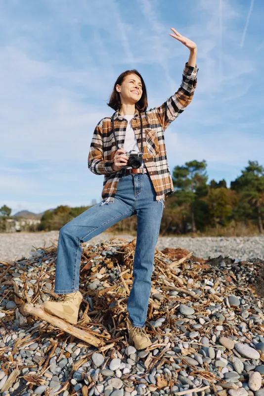 Photographer woman on a rocky beach, casual plaid shirt and denim, holding a camera and raising a hand toward the bright sky in a sunny outdoor shoot.