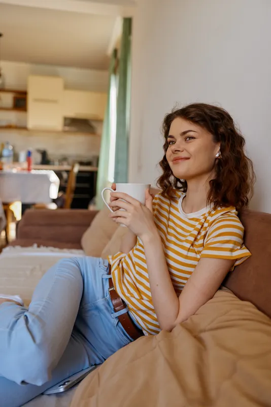 Smiling woman relaxing on a couch at home, holding a mug. Casual striped shirt, jeans, warm light, cozy living room mood. Focus on lifestyle, comfort, wellbeing and everyday routine.
