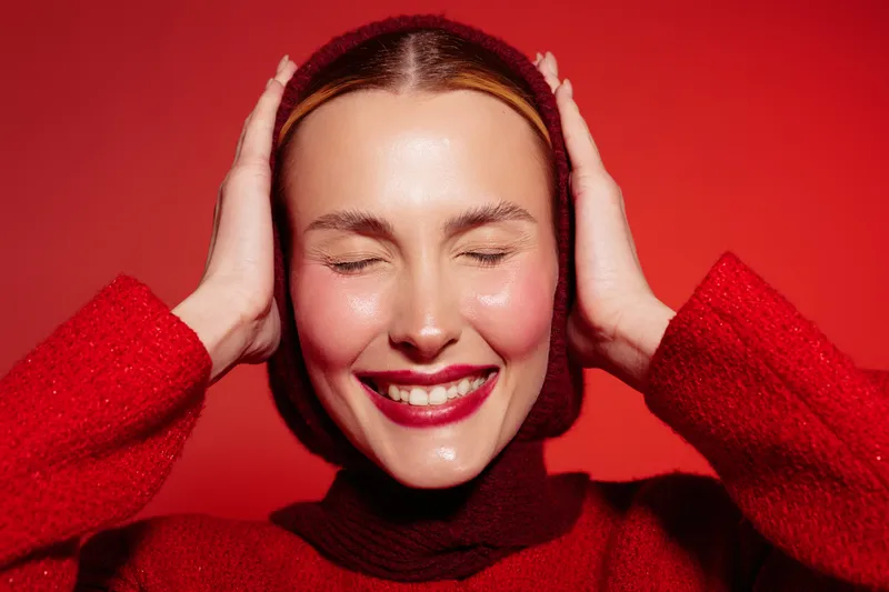 Smiling woman in red sweater with closed eyes holds head with hands on red background. Concept of happiness, joy, and positive emotions in studio portrait.
