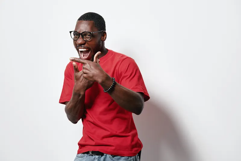 African American man with dark skin wearing glasses and red t shirt laughing and pointing finger gesture isolated on white background with casual style and joyful expression.