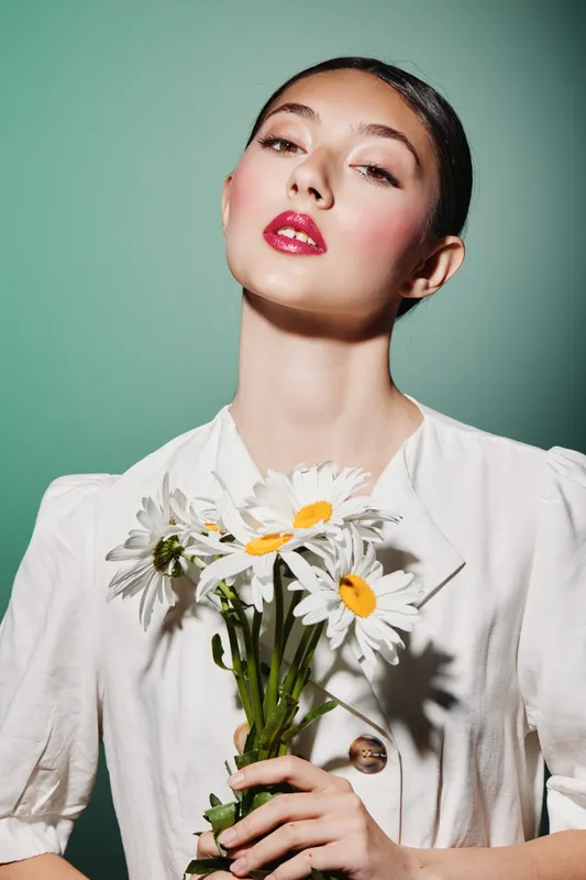 Elegant woman with glossy red lips holding chamomile flowers looks confidently at camera on olive background. Concept of beauty, fashion headline creative portrait lighting.