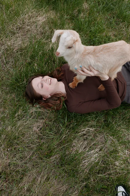 Relaxed woman lying on grass plays with a young goat kid, enjoying outdoor time, gentle bonding, casual lifestyle, sunny farm vibe, wholesome moment