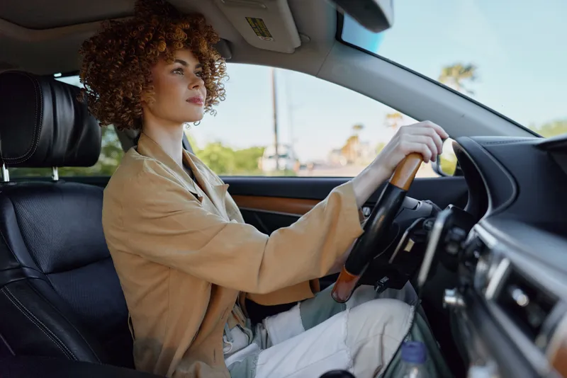 Cheerful woman driving a car, sunlit interior, warm tones, relaxed posture, curly hair, casual jacket, open road glimpse, soft breeze, natural colors, lifestyle moment