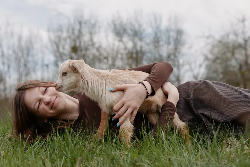 Young woman lying on grass embraces a baby goat outdoors in a natural setting during spring, showing affection and joy in a relaxed lifestyle moment.