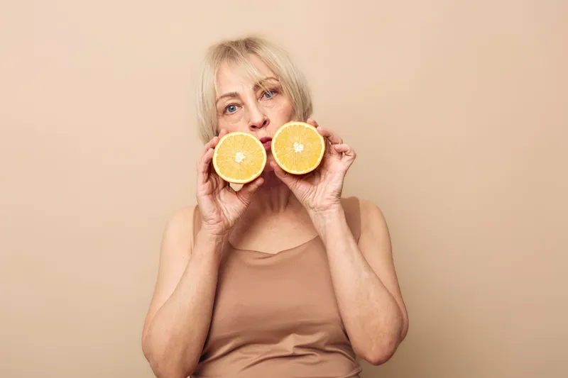 Mature woman with short blonde hair holds two halves of an orange in front of her face against a beige background. Concept of healthy lifestyle, natural nutrition, and freshness.
