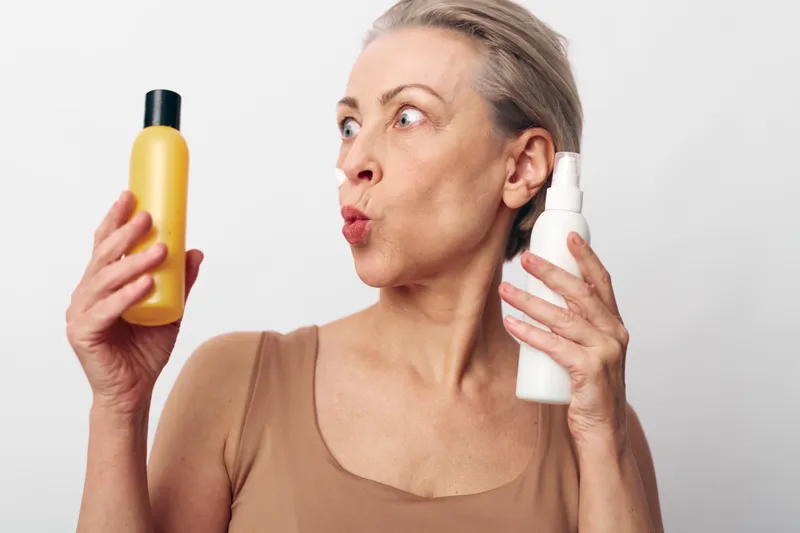 Mature woman with short hair holds yellow bottle and white spray, choosing between skincare products, isolated on white background. Concept for beauty routine and personal care decision.