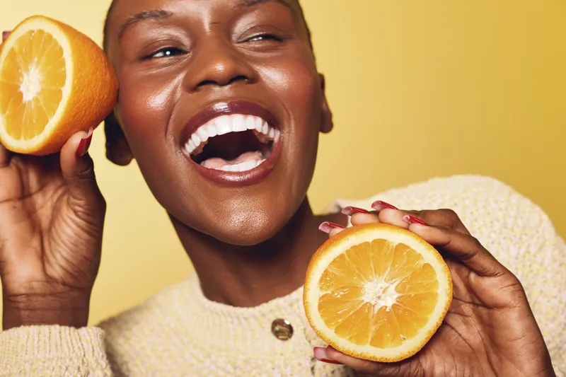 Happy African woman with dark skin holding two orange halves near face, smiling with open mouth on yellow background. Concept of healthy lifestyle, fruit, joy, and natural beauty.