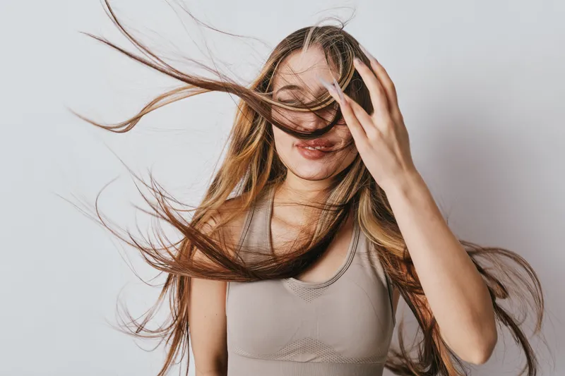 Young woman with long hair blowing in wind and hand touching face wearing sleeveless top on light background. Portrait with motion and natural expression.