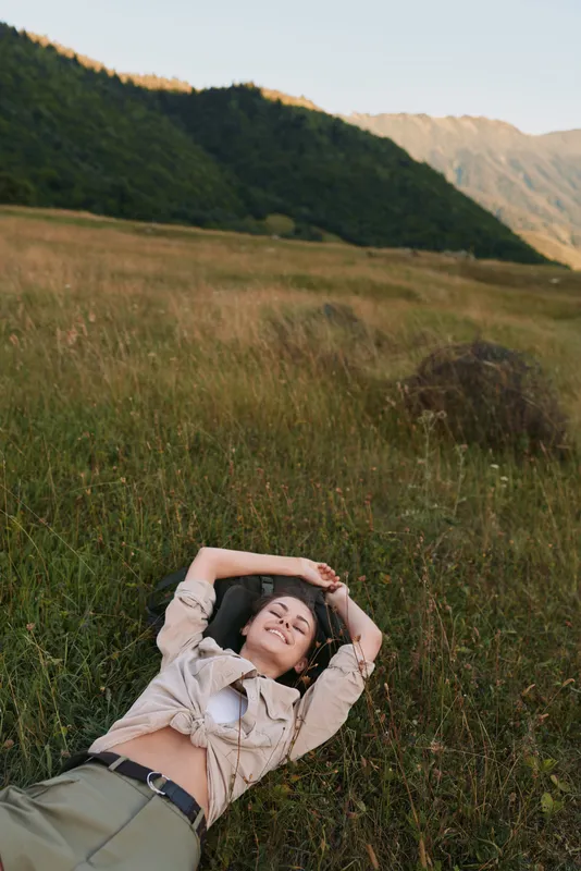 A woman lies in a grassy meadow, arms folded behind her head, smiling softly. Broad hills and warm light create a serene outdoor lifestyle portrait today
