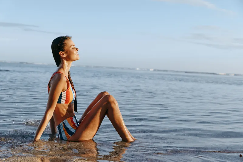 Young woman in a striped bikini sits at the edge of the calm sea, eyes closed, basking in warm sun and a gentle breeze, relaxed and content.