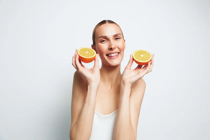 Smiling woman holding two halves of fresh orange near face, bright studio lighting, clean white background, healthy lifestyle concept, natural beauty expression