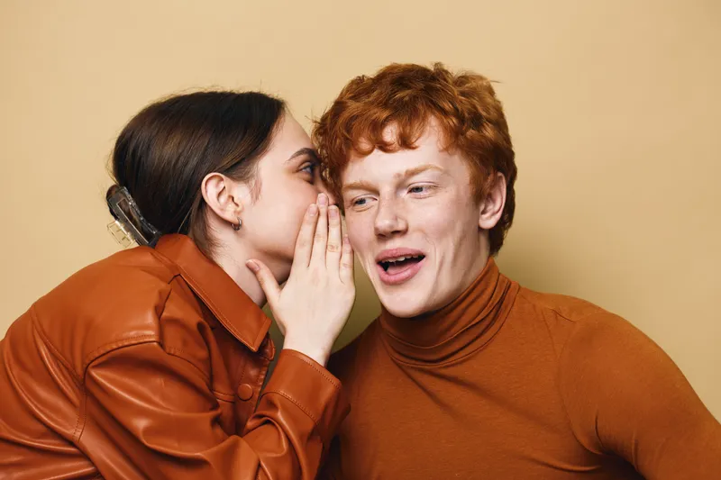Young woman whispering secret to young man wearing brown turtleneck and jacket on beige background. Closeup, communication, friendship and expression of surprise and trust.
