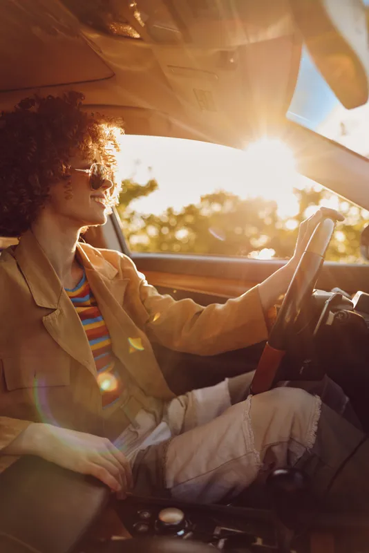 Young woman with curly hair drives a car at sunset, wearing a tan jacket and striped top, capturing a retro, laid back mood.