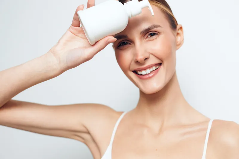 Smiling woman holding white cosmetic bottle near face, clear skin, natural makeup, bright studio background, skincare concept, beauty routine, healthy lifestyle, closeup portrait