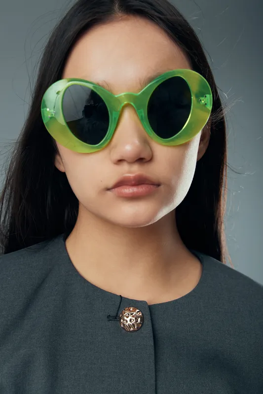 Young woman wearing oversized green sunglasses and dark gray clothing posing in front of neutral gray background with serious expression and long dark hair visible.